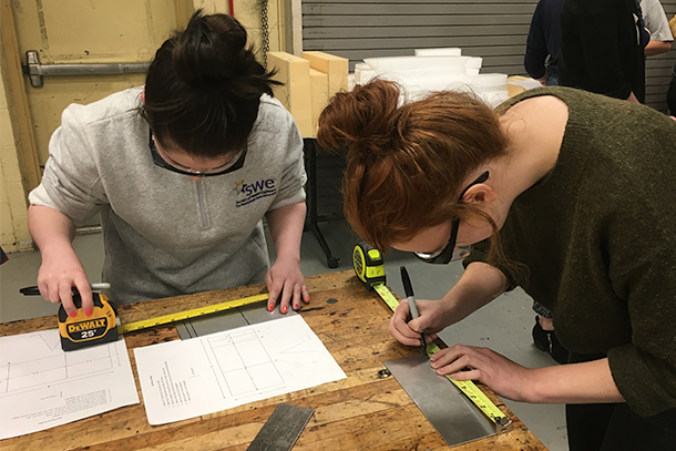 Two engineering students measure and mark sheet metal to create a smartphone holder.