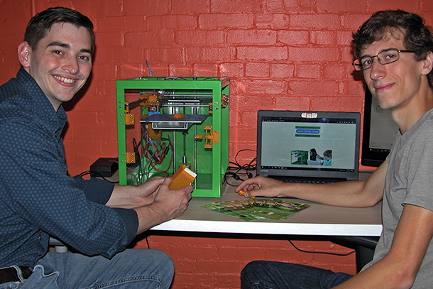 Two men sit by a table that a computer and a 3D printer are on top of.