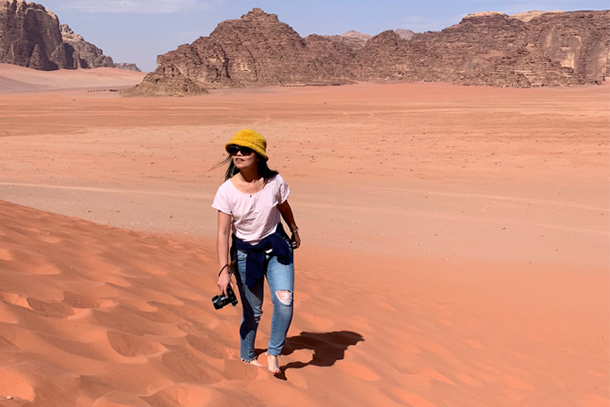 Young woman wearing sunglasses, a yellow hat, white t-shirt and jeans walks through the desert with a camera and stares off into the distance.